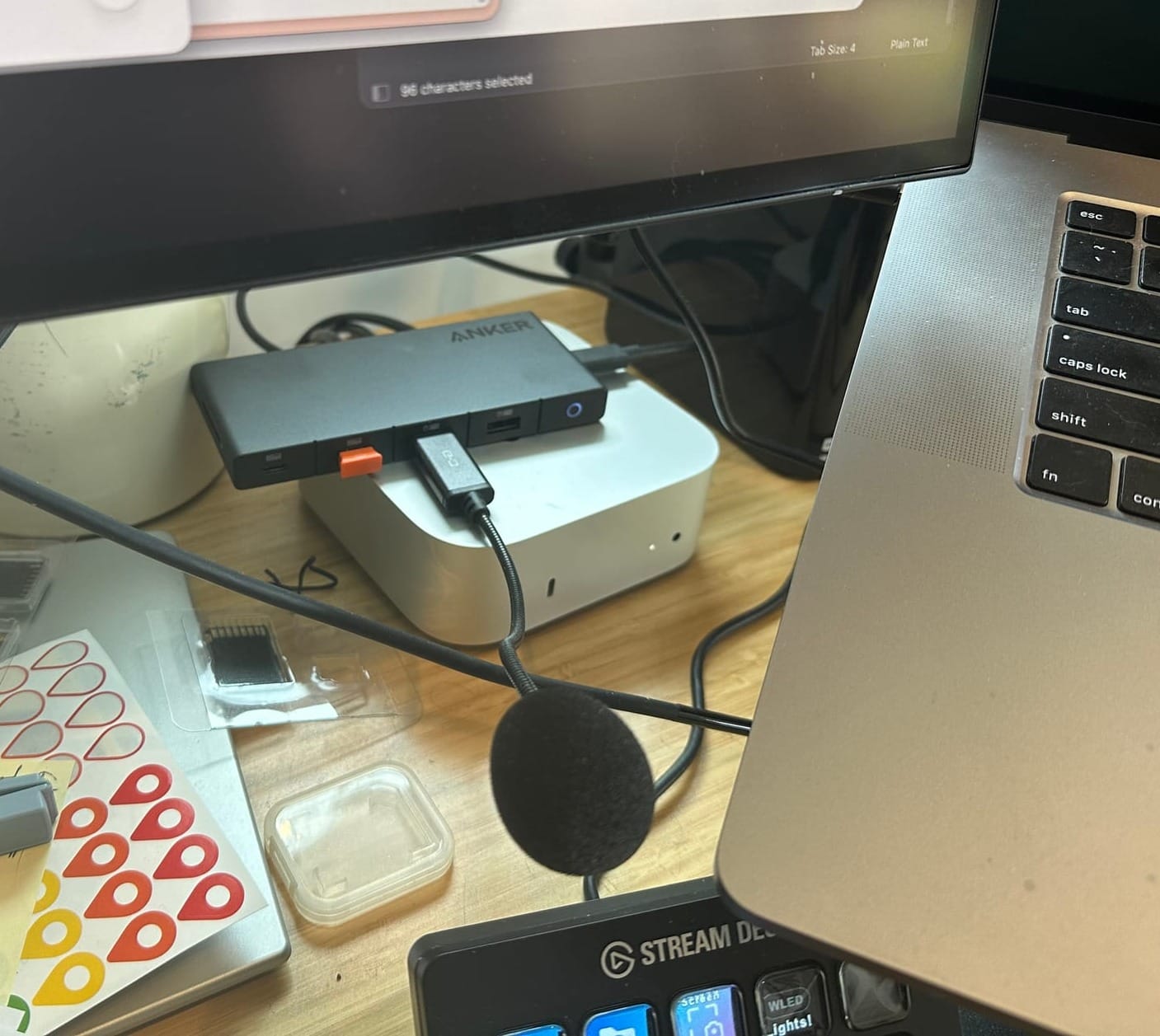 A cluttered desk workspace showing the setup where JAWS AI agent operates, featuring a curved monitor displaying a web interface, an Anker USB hub, Stream Deck controller, and various cables connecting a MacBook Pro to external devices on a wooden surface.